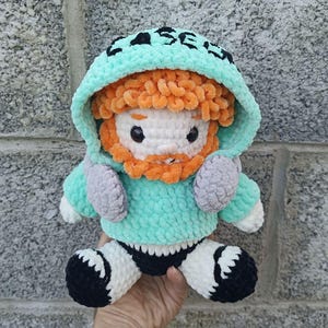 Könnte beinhalten: Handgemachte Häkelpuppe mit orangefarbenen Haaren und Bart, trägt einen türkisfarbenen Kapuzenpullover mit dem Wort "SEAS" auf der Kapuze. Die Puppe hat graue Fäustlinge, eine schwarz-weiß gestreifte Hose und weiße Schuhe.