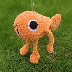 Puede incluir: Un juguete de pez naranja de crochet con ojos negros y pupilas blancas. El pez tiene dos patas largas de color naranja y una aleta de cola. El juguete está sentado en un lecho de hierba verde.