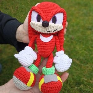 May include: A handmade crochet Knuckles the Echidna plush toy. The red character has white gloves, a white crescent on its chest, and large red shoes with yellow stripes. The plush is held in a person's hands against a green background.