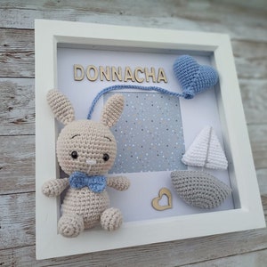 Può includere: Una scatola ombra con cornice bianca con un coniglietto all'uncinetto con papillon blu, un cuore blu e una barca a vela. Il nome "DONNACHA" è scritto con lettere di legno sopra uno spazio fotografico con uno sfondo a pois blu e bianco.