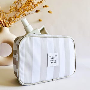 Puede incluir: Bolsa de cosm&eacute;ticos rectangular a rayas grises y blancas, con cremallera blanca y una peque&ntilde;a etiqueta que dice "consume less create more". Una botella blanca es parcialmente visible dentro de la bolsa. Un jarr&oacute;n beige con plantas secas est&aacute; en el fondo.