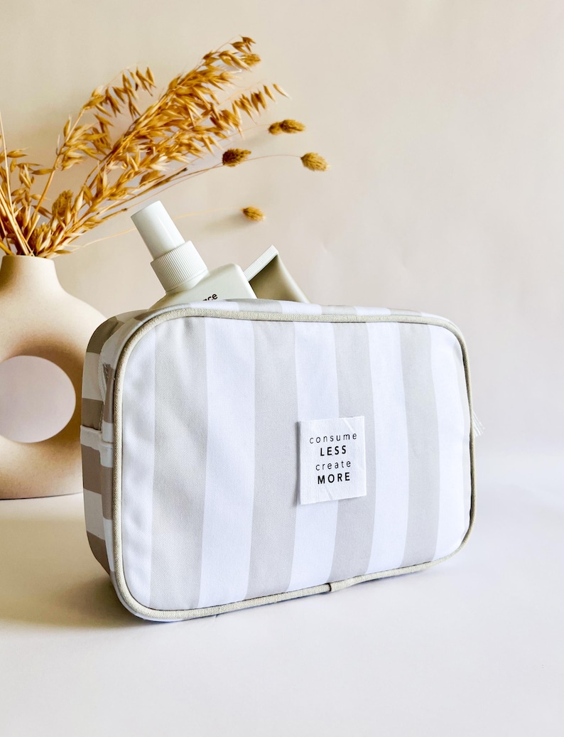 Puede incluir: Bolsa de cosm&eacute;ticos rectangular a rayas grises y blancas, con cremallera blanca y una peque&ntilde;a etiqueta que dice "consume less create more". Una botella blanca es parcialmente visible dentro de la bolsa. Un jarr&oacute;n beige con plantas secas est&aacute; en el fondo.