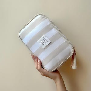 Puede incluir: Un neceser rectangular con rayas horizontales beige y blancas. La bolsa tiene una cremallera color crema y una borla. Una peque&ntilde;a etiqueta blanca en la parte delantera dice "Consume Less Create More".
