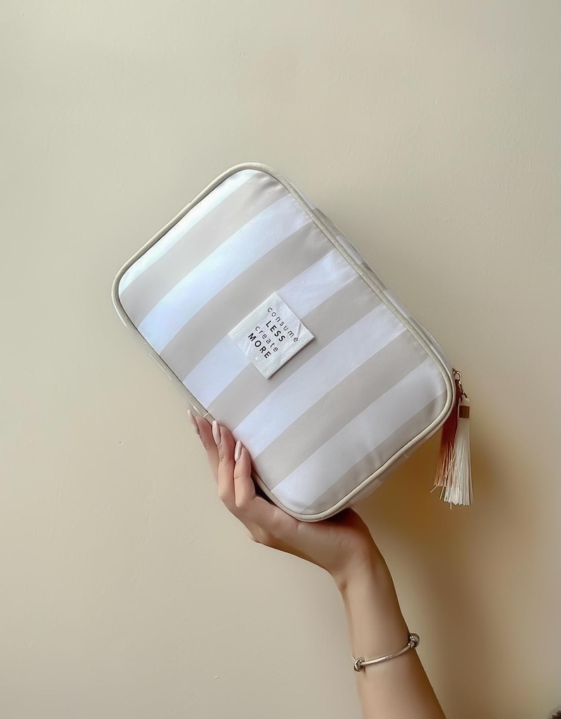 Puede incluir: Un neceser rectangular con rayas horizontales beige y blancas. La bolsa tiene una cremallera color crema y una borla. Una peque&ntilde;a etiqueta blanca en la parte delantera dice "Consume Less Create More".