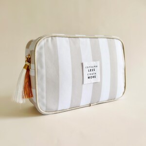 Puede incluir: Un neceser rectangular con rayas verticales grises y blancas. La bolsa tiene una cremallera dorada y una borla con hilos blancos y naranjas. Una peque&ntilde;a etiqueta blanca en la bolsa dice "consume less create more."