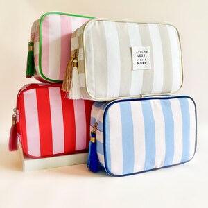 Puede incluir: Cuatro neceseres rectangulares en rosa, rojo, gris y azul con rayas verticales. Cada bolsa tiene una borla a juego y una cremallera dorada. La bolsa gris tiene una etiqueta blanca que dice "consume less create more."