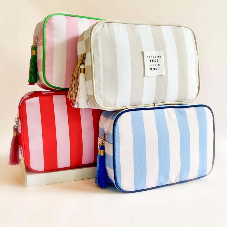 Puede incluir: Cuatro neceseres rectangulares en rosa, rojo, gris y azul con rayas verticales. Cada bolsa tiene una borla a juego y una cremallera dorada. La bolsa gris tiene una etiqueta blanca que dice "consume less create more."