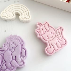 May include: A collection of pastel-colored, whimsical soap shapes. The assortment includes a unicorn, a rainbow, a castle, and a horse. The soaps are in shades of pink, purple, and white, presented in a white box and scattered on a white surface.