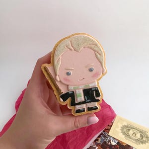 May include: A decorated cookie in the shape of a cartoon character with blonde hair, wearing a black robe and holding a wand. The cookie has pink skin and is held in a hand. The cookie is on a white background with a red tissue paper and some cards.
