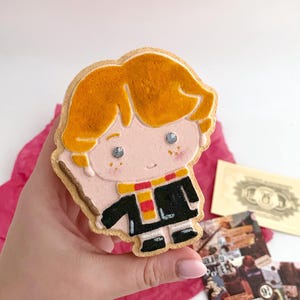 Pode incluir: Um biscoito decorado em forma de personagem de desenho animado com cabelo laranja, vestindo uma túnica preta e um cachecol amarelo e vermelho. O biscoito é segurado em uma mão, com um papel vermelho e outros itens no fundo.