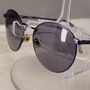 May include: Round sunglasses featuring a dark purple metal frame and dark tinted lenses. The eyewear includes thin metal temples and gold-toned nose pads. The design is characterized by a circular lens shape and a slim bridge connecting the two rims.