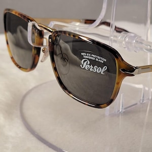 May include: Tortoiseshell-patterned sunglasses with dark grey lenses and gold-tone metal temples. The right lens features a white sticker with the text Persol, 100% U.V. PROTECTION, and TEMPERED GLASS. The frames have a classic rectangular shape.