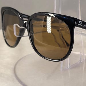 May include: Black sunglasses with brown tinted lenses and a glossy finish. The temple arm features the white printed text RL 2000. The frames have a rounded shape with a bridge connecting the two lenses, providing a classic look for eyewear.