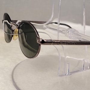 May include: Round sunglasses with a dark metallic gunmetal frame and dark green lenses. The temples feature a textured, ribbed pattern along the sides, and the earpieces are finished with black tips. The bridge includes clear nose pads.