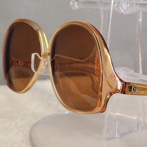Puede incluir: Gafas de sol de gran tamaño con montura de plástico color ámbar translúcido y lentes marrones degradadas. Las patillas presentan un pequeño detalle metálico en tono plateado con una letra e minúscula grabada cerca de la bisagra.