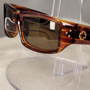 May include: Tortoiseshell sunglasses with rectangular frames and dark brown lenses. The temples feature a gold-colored cross emblem and three small gold-colored studs near the hinge. The frame has a glossy finish with a distinct brown and amber pattern.