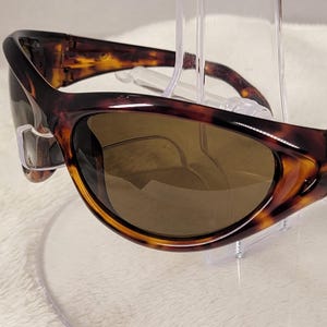 May include: Tortoiseshell sunglasses with a wrap-around frame design and dark brown tinted lenses. The temples feature the Spy brand logo embossed on the side. These sunglasses have a curved, aerodynamic shape with a glossy finish throughout.
