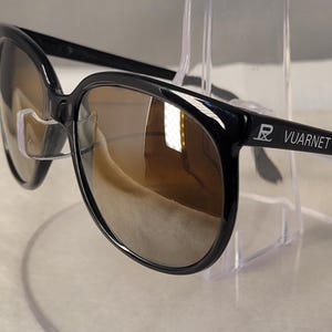 May include: Black sunglasses with rounded frames and brown tinted lenses. The temples feature the white Vuarnet logo and the brand's signature ski icon. These glasses have clear nose pads and a glossy finish, showcasing a classic design.