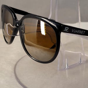 May include: Black Vuarnet sunglasses featuring a rounded frame and brown tinted lenses. The temple arm displays the Vuarnet brand name and logo in white lettering. These sunglasses have curved earpieces and clear nose pads for support.