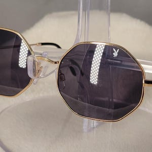 May include: Octagonal sunglasses featuring thin gold-tone metal frames and dark gray tinted lenses. The right lens displays a small white rabbit logo. The temples are thin gold metal with black plastic tips, and the bridge includes clear nose pads.