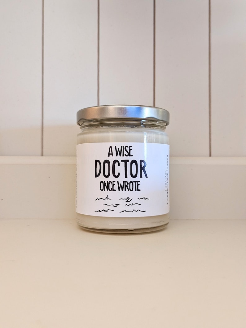 Puede incluir: Un tarro de vela blanco con tapa plateada. La etiqueta dice "A Wise Doctor Once Wrote" con una serie de l&iacute;neas onduladas debajo.