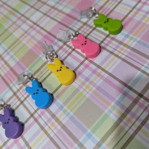 May include: Six colorful bunny-shaped charms with a white background. The charms are yellow, pink, green, blue, and purple. The charms are attached to clear plastic strings.