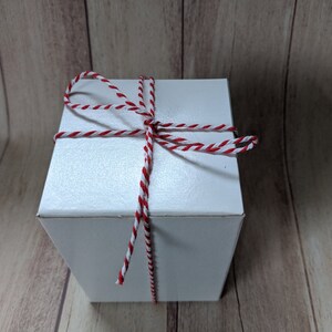 Puede incluir: Una caja de regalo blanca atada con cuerda de panadero roja y blanca.