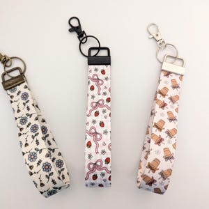 May include: Three fabric wrist keychains with different patterns. One has blue flowers, another has pink bows and ladybugs, and the third has ice cream cart illustrations. Each keychain has a metal clasp.