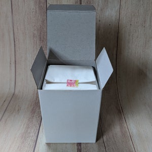 Puede incluir: Una caja de cart&oacute;n blanca con una tapa gris est&aacute; abierta, revelando una bolsa de papel de seda blanca con una pegatina de dise&ntilde;o floral rosa y amarillo. La caja est&aacute; sentada sobre una superficie de madera.