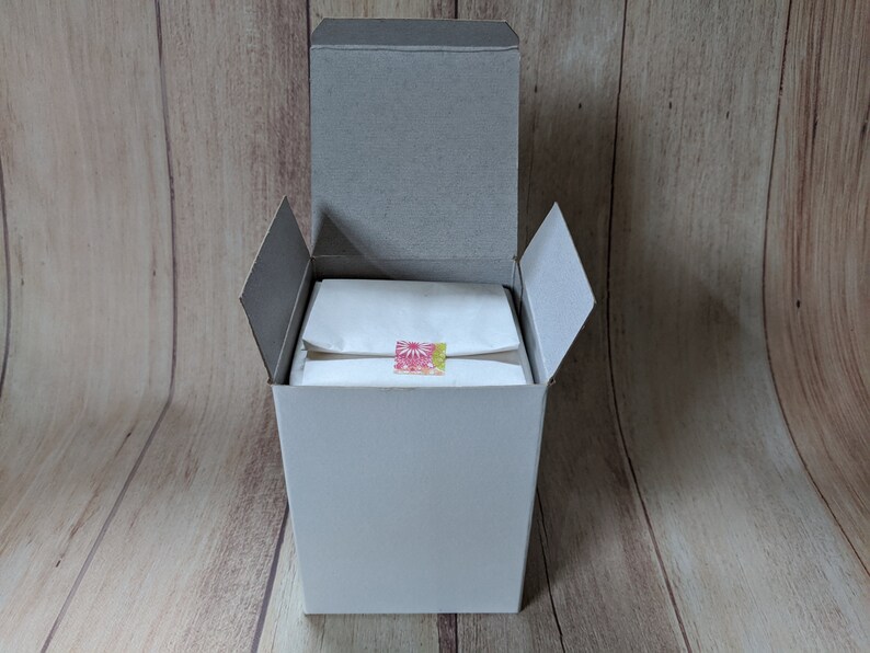 Puede incluir: Una caja de cart&oacute;n blanca con una tapa gris est&aacute; abierta, revelando una bolsa de papel de seda blanca con una pegatina de dise&ntilde;o floral rosa y amarillo. La caja est&aacute; sentada sobre una superficie de madera.