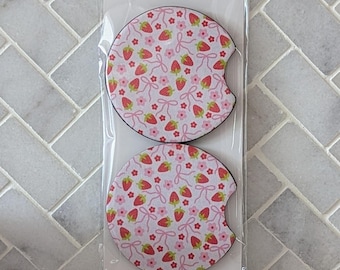 May include: Two car coasters with a strawberry pattern. The coasters are round with a curved cutout for easy removal. The strawberry pattern includes red strawberries, pink flowers, and bows on a white background. The packaging reads "Car Coasters" and "Handmade with love."