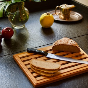 Puede incluir: Una tabla de cortar de madera con un cuchillo para pan y rebanadas de pan. La tabla tiene un diseño acanalado y está hecha de madera marrón claro. El cuchillo tiene un mango negro y una hoja plateada.