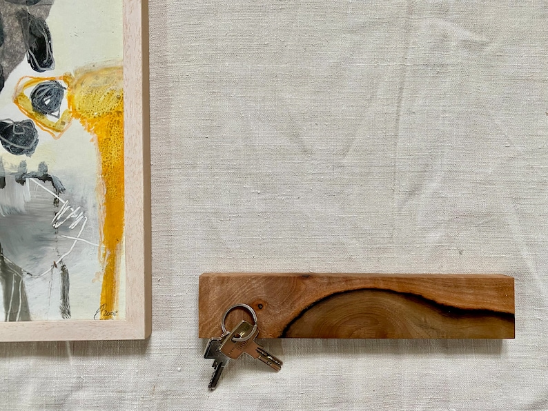 Pu&ograve; includere: Un porta chiavi in legno con la parte superiore curva e un set di chiavi appese. Il porta chiavi &egrave; su una superficie beige chiaro.