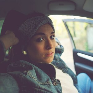 May include: A woman wearing a gray knitted headband and a denim jacket sits in the driver's seat of a car. She is looking out the window.