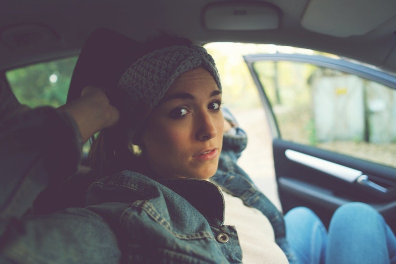 May include: A woman wearing a gray knitted headband and a denim jacket sits in the driver's seat of a car. She is looking out the window.