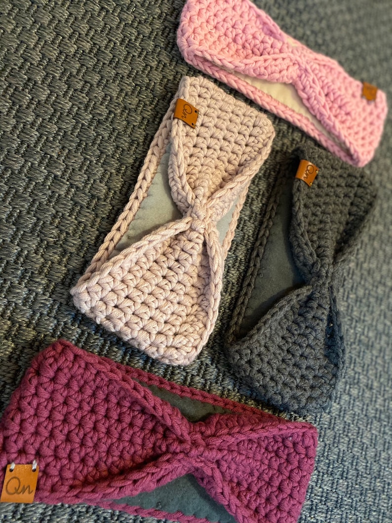 May include: Three crocheted headbands in pink, gray, and burgundy with a brown leather tag that says "Qn". Each headband has a soft, fleece lining.