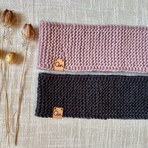 Könnte beinhalten: Zwei gestrickte Stirnbänder, eines in Staubrosa und eines in Anthrazitgrau, werden auf einer neutralen Oberfläche gezeigt. Jedes Stirnband hat ein kleines, quadratisches Lederetikett mit den Buchstaben "Qn". Die Stirnbänder sind aus dickem, strukturiertem Garn gefertigt.