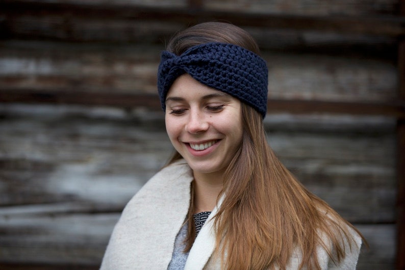 May include: A woman wearing a navy blue crocheted headband with a knot on top. She is smiling and looking down. She is wearing a white sweater.