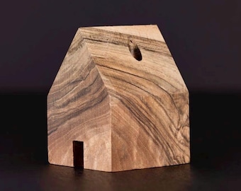 Può includere: Un piccolo oggetto di legno a forma di casa, realizzato in legno scuro con una venatura. La casa ha un piccolo foro nella parte superiore e un'apertura rettangolare nella parte anteriore.