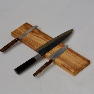 Puede incluir: Tres cuchillos de cocina sobre una tabla de madera. Los cuchillos tienen hojas de acero inoxidable y mangos de madera. El cuchillo más grande tiene un mango negro. La tabla es de madera marrón claro.