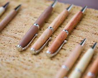 Può includere: Una collezione di penne in legno con dettagli argentati. Le penne variano di colore, mostrando la venatura naturale del legno, dal beige chiaro al marrone scuro. Sono disposte su una superficie strutturata marrone chiaro.