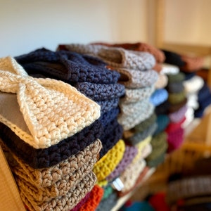 May include: A stack of crocheted headbands in various colors, including beige, brown, dark blue, and light blue. The headbands are arranged on a wooden shelf.