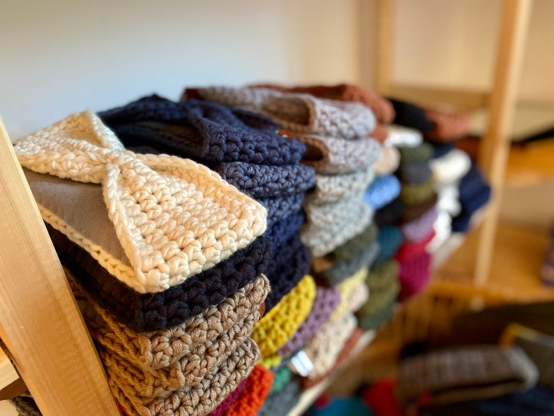 May include: A stack of crocheted headbands in various colors, including beige, brown, dark blue, and light blue. The headbands are arranged on a wooden shelf.