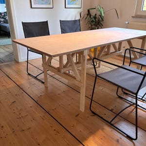 Peut inclure: Une table à manger en bois clair avec un plateau rectangulaire et un piètement simple à cadre ouvert. La table est entourée de quatre chaises en métal noir avec des sièges en tissu gris.