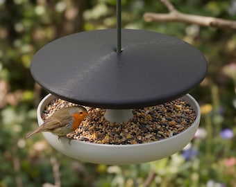 May include: A bird feeder with a black top and a white bowl filled with birdseed. A small bird with orange and brown feathers is perched on the edge of the bowl, eating. The feeder is suspended by a thin metal rod.