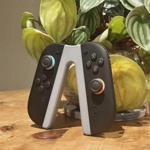 以下が含まれることがあります： 青とオレンジのジョイスティックが付いた、黒とグレーのゲームコントローラー。 コントローラーは、両側を接続するライトグレーのブリッジを備えたユニークなデザインです。 ボタンには文字と記号がマークされています。