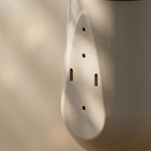 May include: Close-up of a light gray, cylindrical object with a series of small holes and slots. The object has a rounded shape and a smooth surface. The background is blurred, with hints of light and shadow.