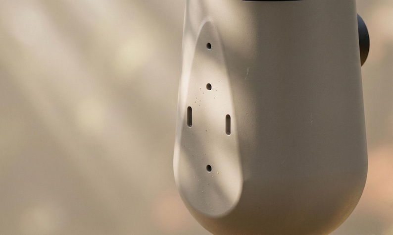 May include: Close-up of a light gray, cylindrical object with a series of small holes and slots. The object has a rounded shape and a smooth surface. The background is blurred, with hints of light and shadow.