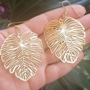 Puede incluir: Un par de pendientes calados dorados con forma de hojas de Monstera. Los pendientes tienen cierre de gancho y se sostienen en la mano de una persona sobre un fondo verde borroso. Los pendientes son un artículo de joyería de temática tropical.