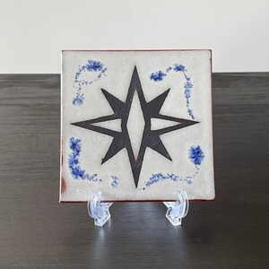 May include: A white ceramic tile with a black eight-pointed star design. The tile has a blue floral border and is displayed on a clear stand.
